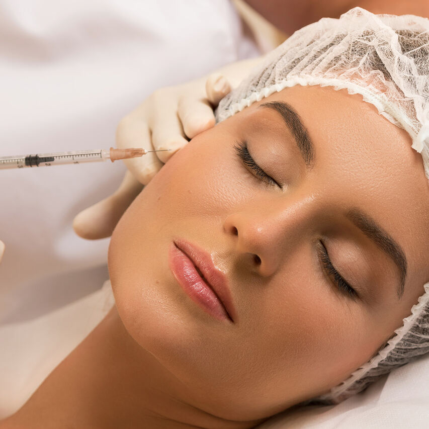 A relaxed woman with closed eyes wearing a white surgical hair cap receives a cosmetic injection from a gloved practitioner holding a syringe near her temple